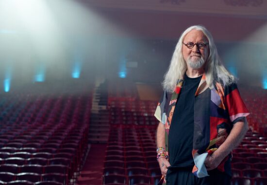 Billy Connolly standing in an empty theatre looking serene.