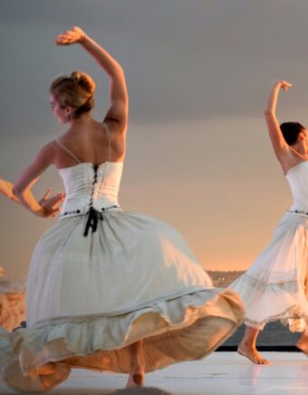 A series of people dance in white dresses on a warm lit stage