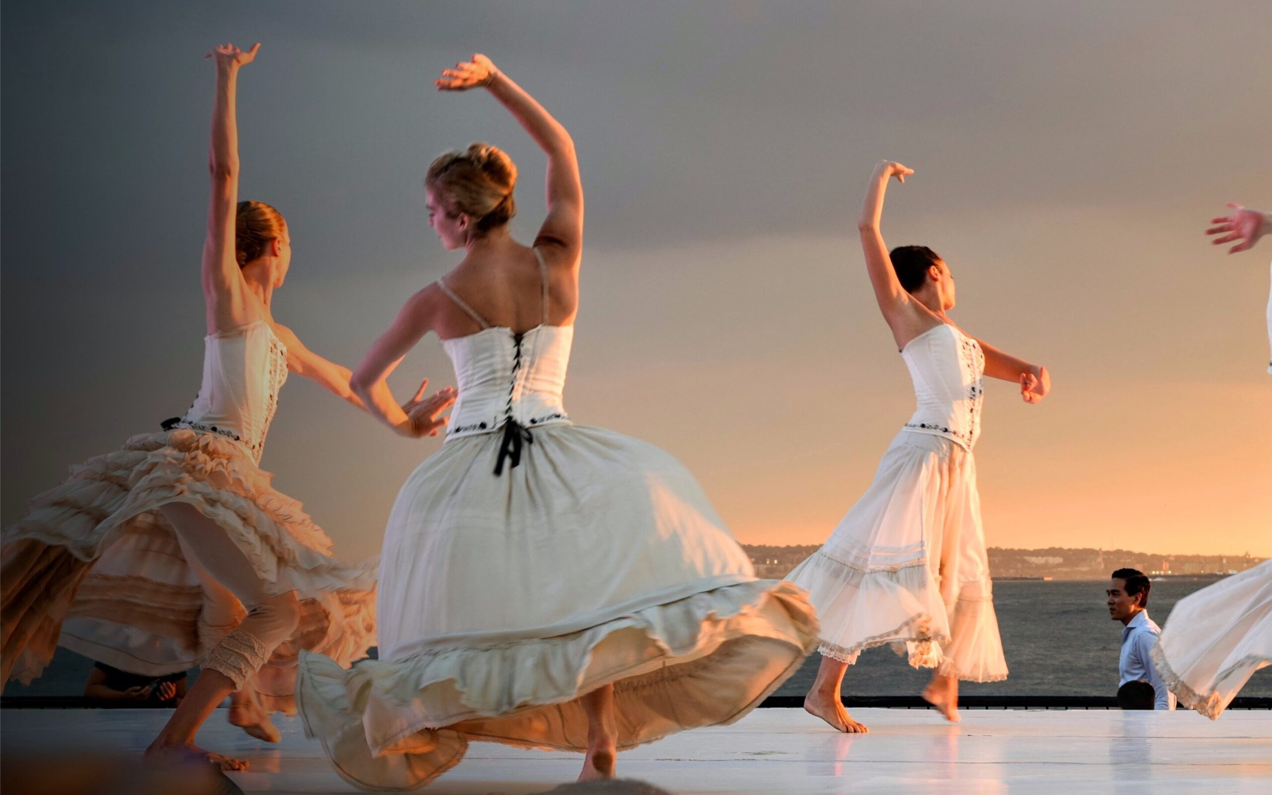 A series of people dance in white dresses on a warm lit stage