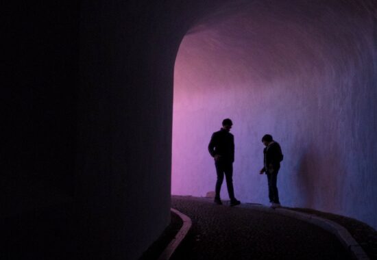 Two figures stand in a dark alleyway, lit by purple light
